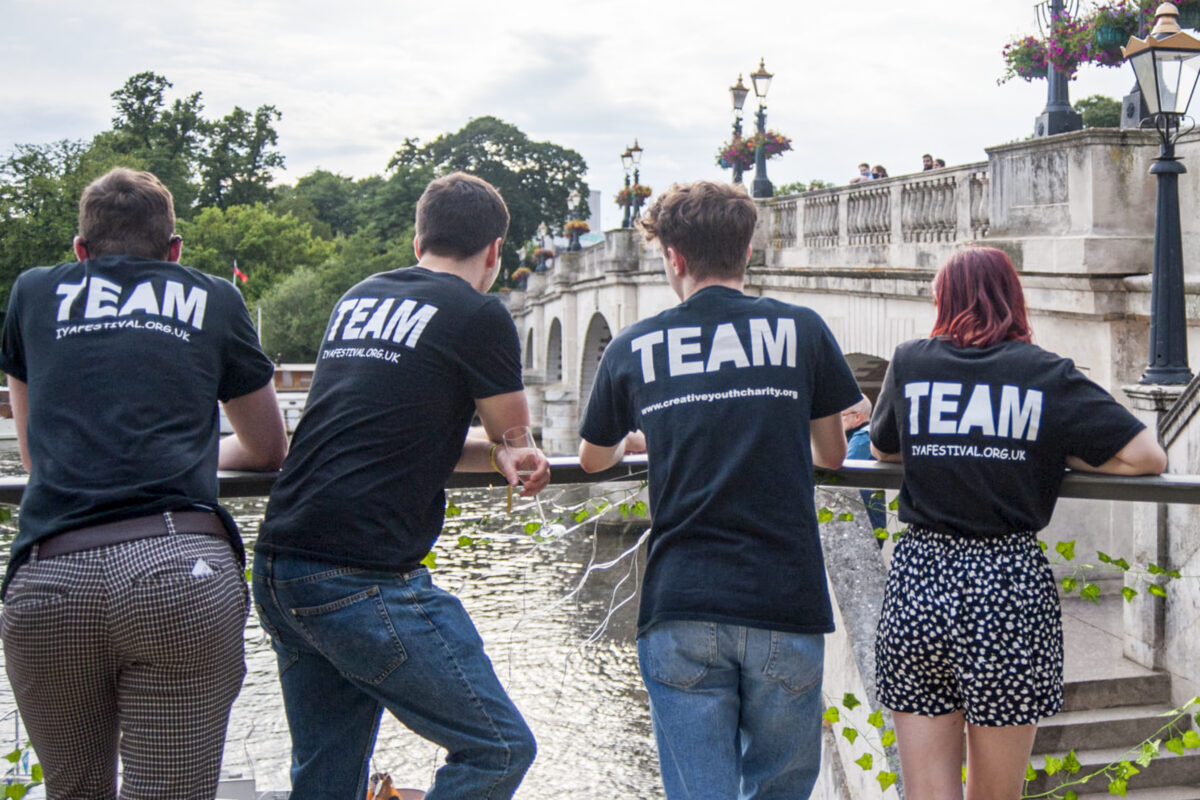 Four young people face away from the camera looking towards Kingston Bridge and the River Thames. They wear black t-shirts which say 'team' on the back.