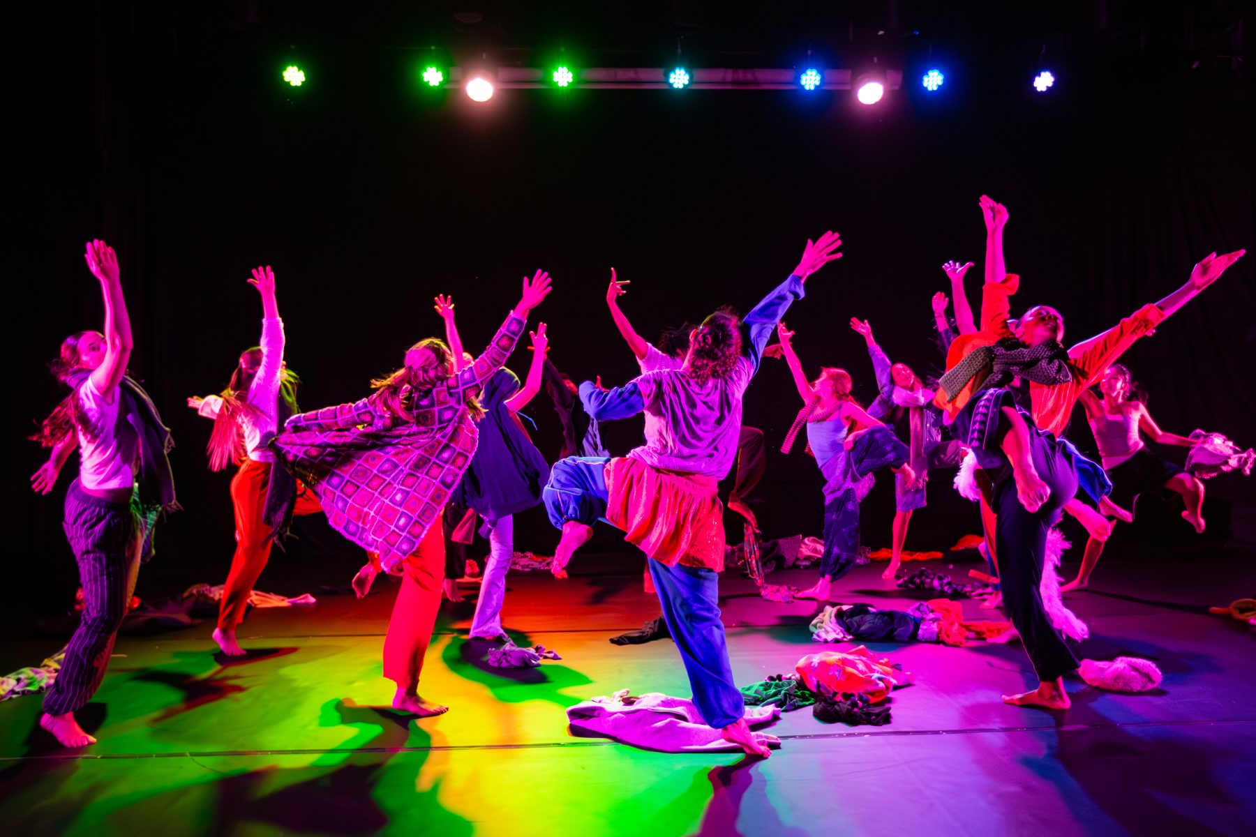 A large group of dancers in purple light hold their arms up in the air, whilst in a circle formation