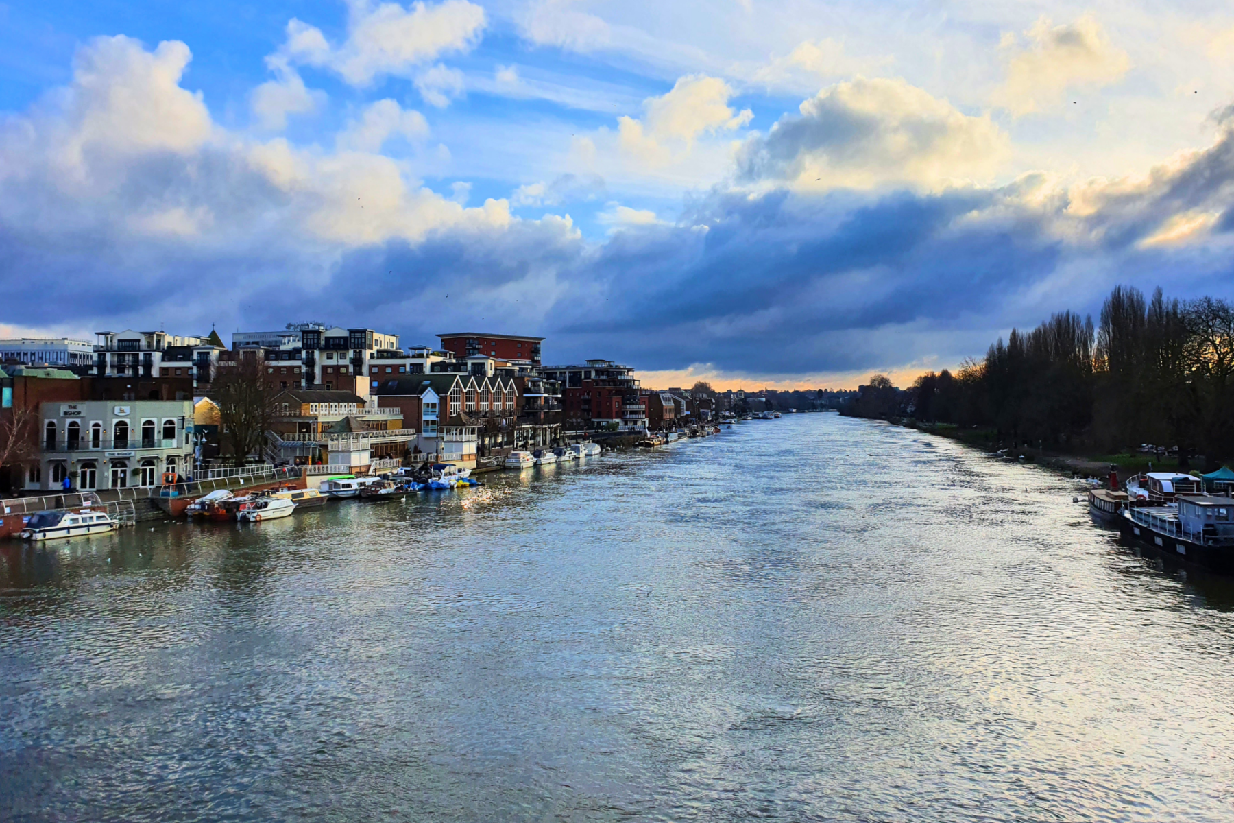 Landscape photo of Kingston's Riverside. Buildings and boats line the left bank, while trees run along the right. The sky is filled with dramatic clouds reflecting light onto the water.
