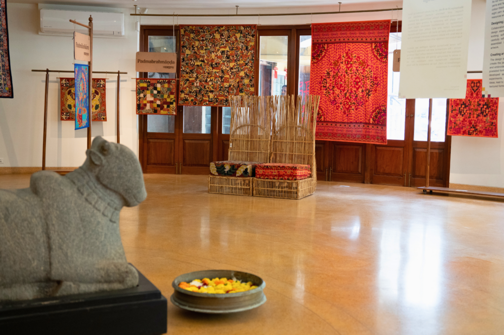 Gallery installation with hanging textile artworks in red, blue, and multicolour geometric patterns displayed on wooden frames and wires. In the foreground, a small stone sculpture of a seated animal faces the exhibition space beside a shallow bowl filled with flower petals. A woven rattan bench with patterned cushions sits centrally, with wooden doors and windows forming the backdrop.