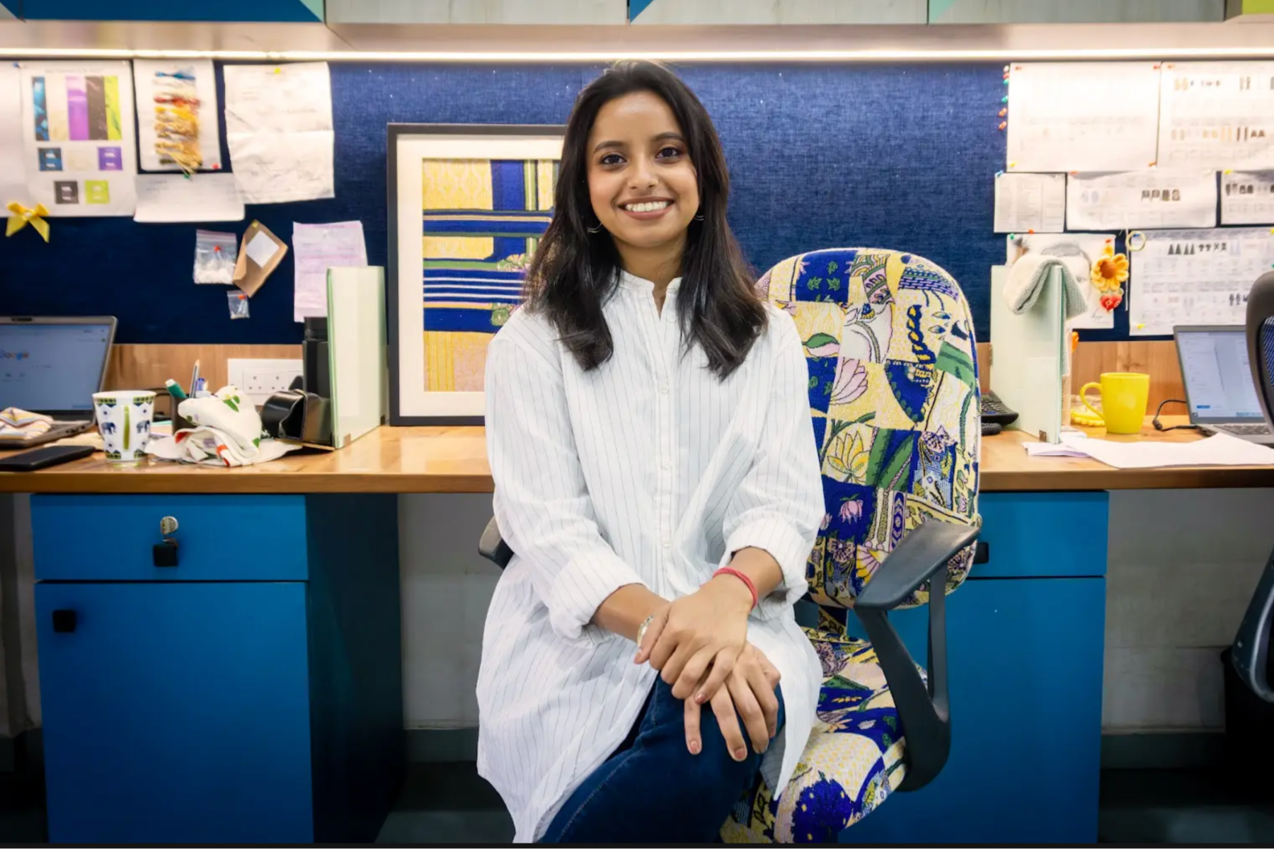 A person sits on a patterned office chair at a desk in a shared workspace, wearing a long white shirt and jeans, with hands resting on one knee; behind them are blue cabinets, a cluttered desk with mugs and stationery, pinned papers on a noticeboard, and framed artwork against a blue wall.